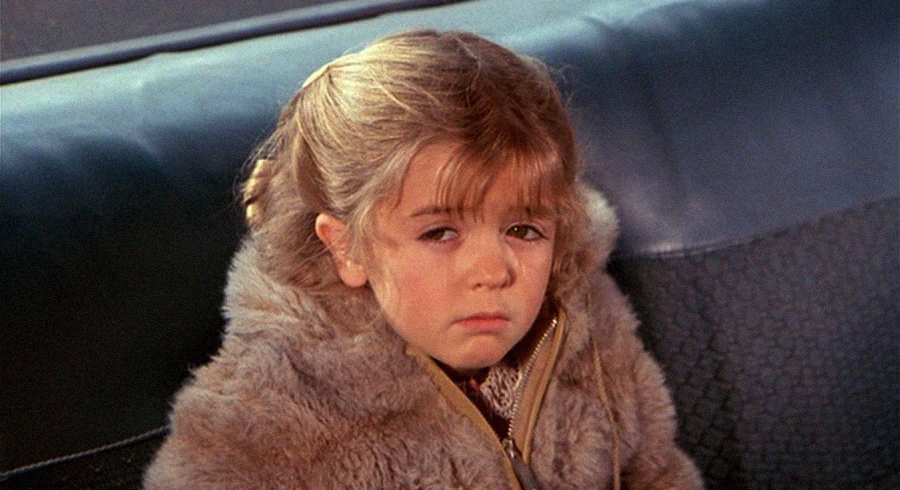 A young girl with blonde hair wearing a fur coat sits on a bench or seat, looking ahead with a tired or sad expression.