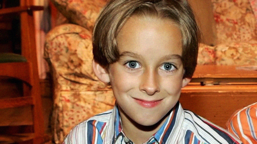 A young child with light brown hair and blue eyes smiles at the camera, wearing a striped shirt. The background features a floral-patterned couch and wooden furniture.