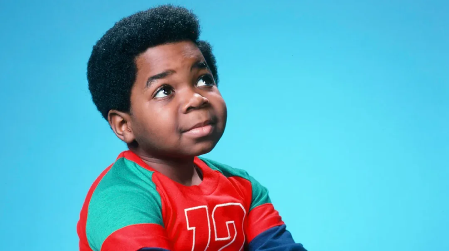 A young boy with short curly hair looks upward with a thoughtful expression. He is wearing a red and green shirt with the number 12 printed on it, and the background is solid light blue.