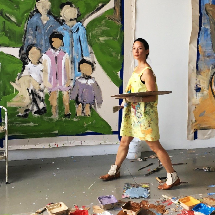 A woman in a paint-splattered dress walks through a studio holding art supplies. Behind her is a large, unfinished painting of four faceless people, with paint trays and brushes scattered on the floor.