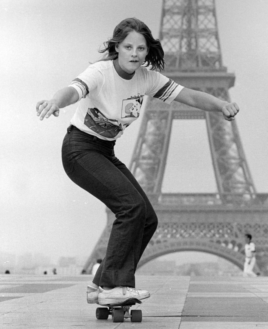 A young woman skateboards in front of the Eiffel Tower, balancing with her arms out while wearing jeans, sneakers, and a printed t-shirt. The scene is in black and white.