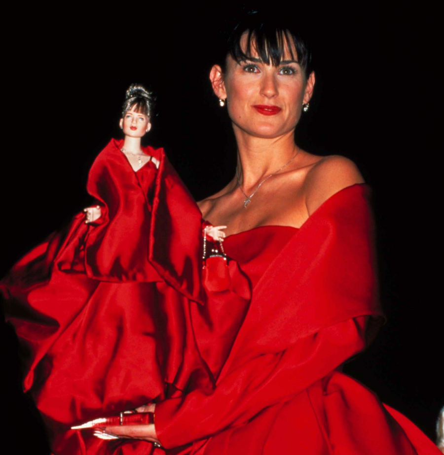 A woman in an elegant off-the-shoulder red gown holds a matching doll dressed in an identical red gown. The background is dark, highlighting both the woman and the doll.
