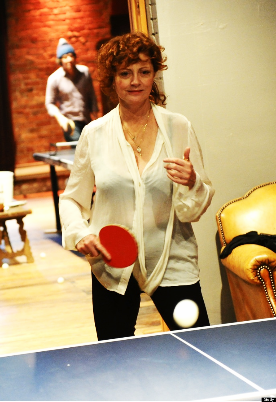 A woman with curly red hair, wearing a white blouse and black pants, plays table tennis indoors. She holds a red paddle and looks focused as she prepares to hit the ball. A man in a blue beanie is in the background near another table.