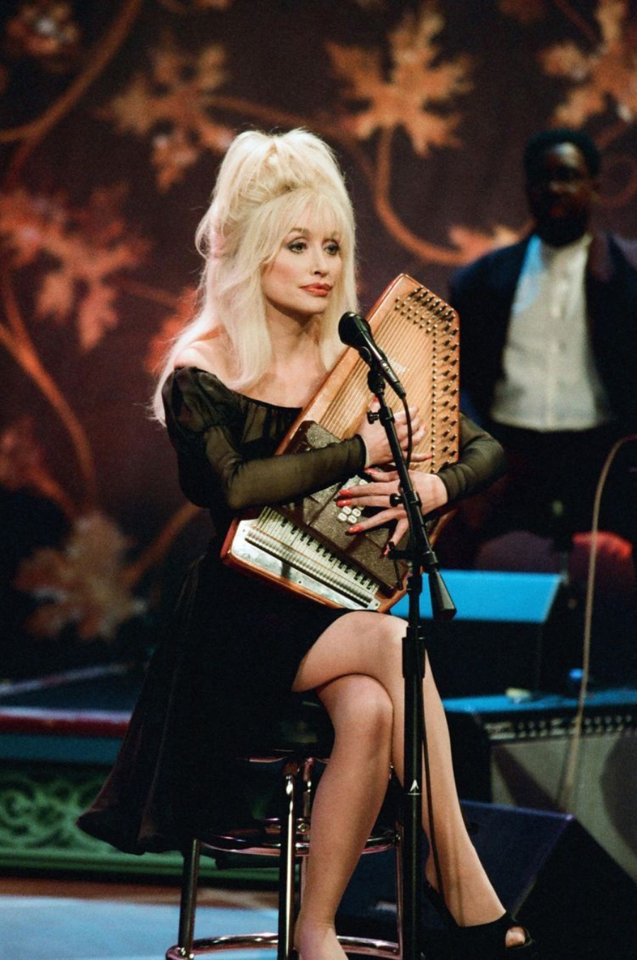 A woman with long blonde hair in a black dress sits on a stool, playing an autoharp and singing into a microphone on stage, with autumn leaf decorations in the background.