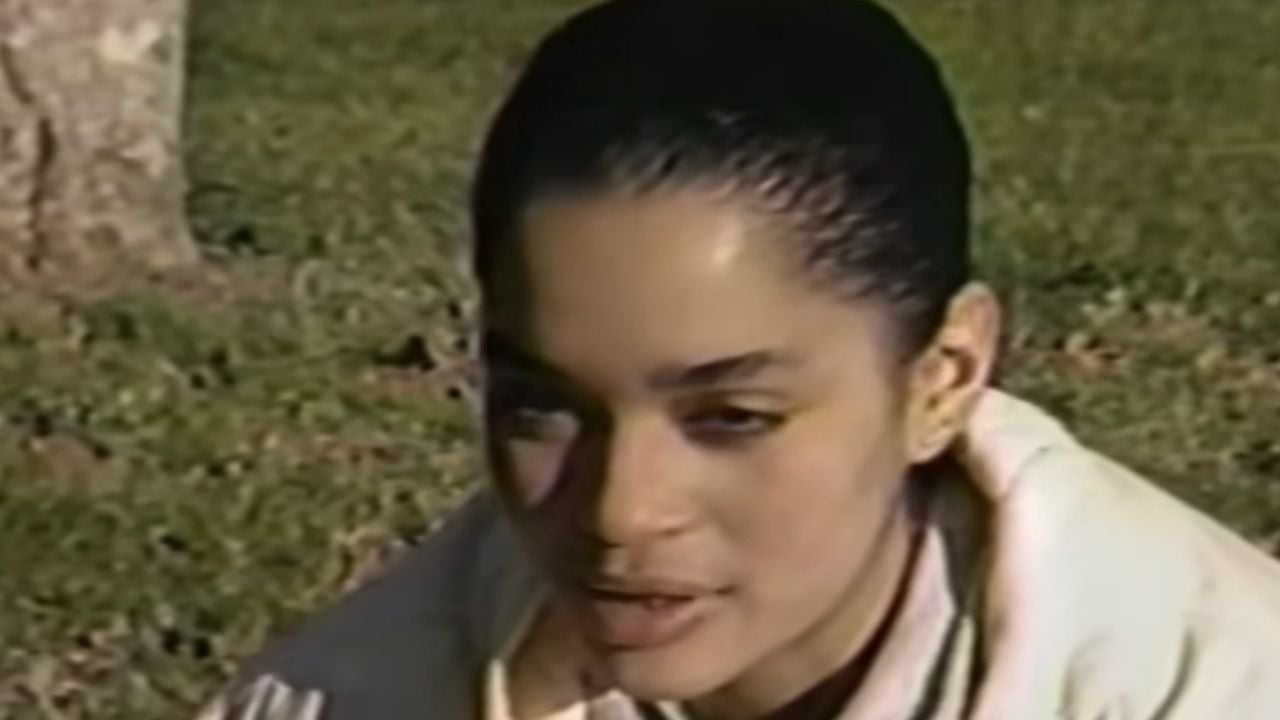 A person with dark hair pulled back is sitting outside on grass, wearing a light-colored jacket. The image has a vintage, slightly grainy quality.