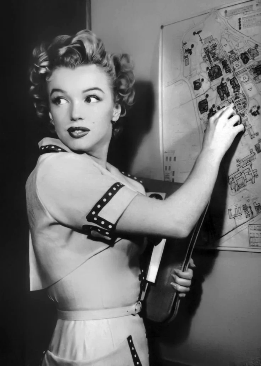 A woman with curled blonde hair and a light-colored dress marks a spot on a large map pinned to a wall, while holding a folder and looking over her shoulder. The image is in black and white.