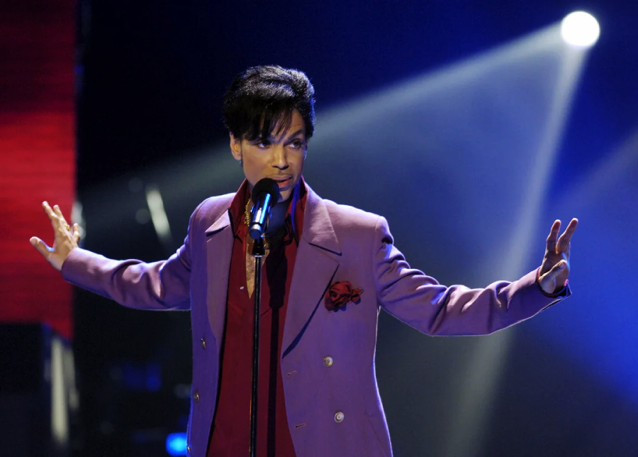 A performer in a purple suit sings into a microphone on stage, spreading their arms wide. Blue and purple stage lights shine behind them, creating a dramatic spotlight effect.
