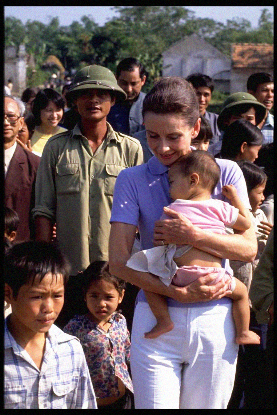 A woman in a light purple shirt gently holds a young child while standing among a group of people, including children and men in green uniforms and helmets, outdoors on a sunny day.