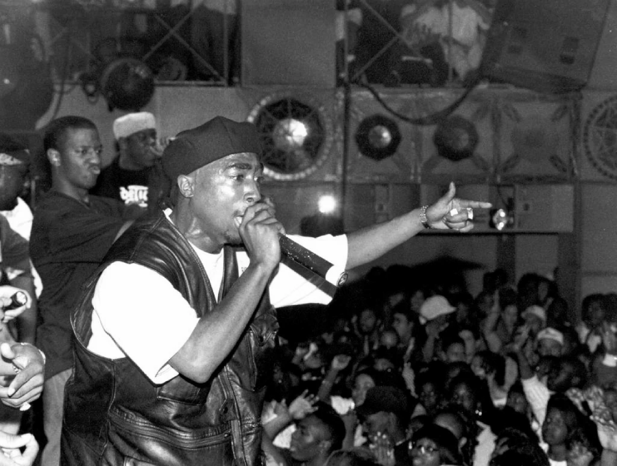 A rapper performs energetically on stage, holding a microphone and pointing toward the crowd. An audience watches, and several people stand behind him. The scene is lively, with fans packed closely together.
