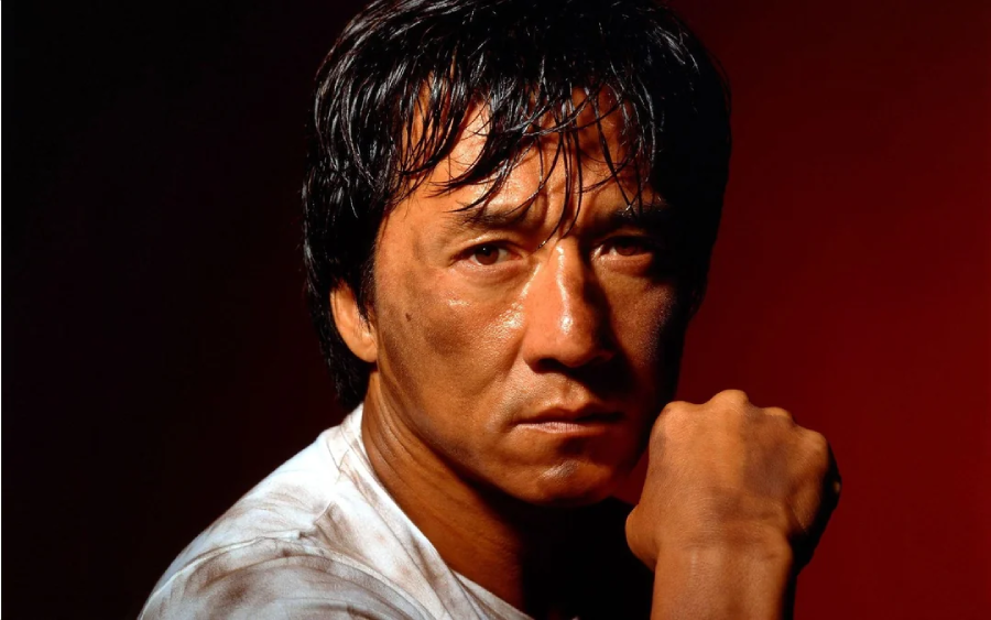 A man with short, dark, wet hair gazes seriously at the camera, resting his chin on his fist. He wears a white shirt and is set against a dark red background. His expression appears intense and thoughtful.