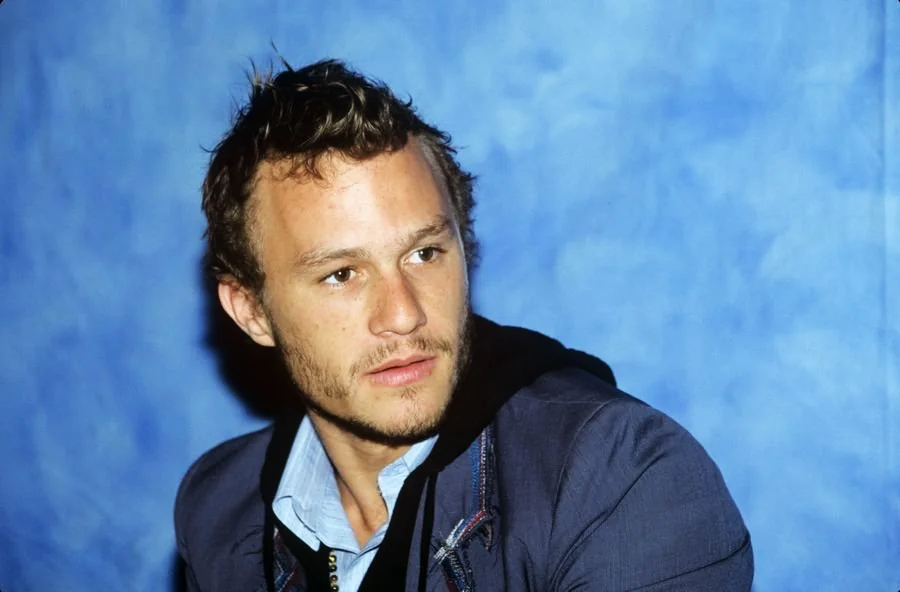 A man with short, tousled hair and a light beard is wearing a blue collared shirt and a dark jacket. He is sitting against a blue, textured background, looking slightly to the side with a thoughtful expression.