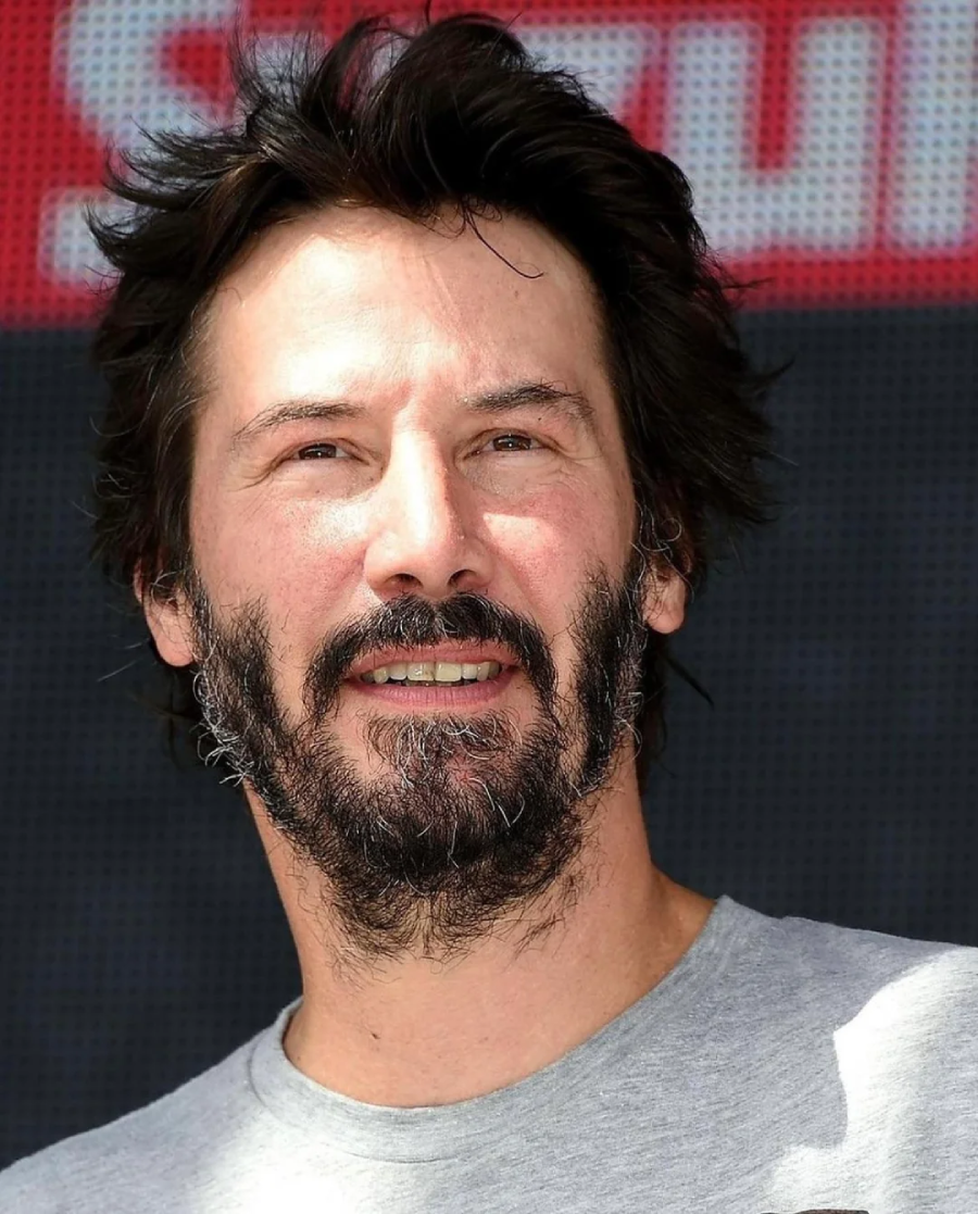 A man with tousled dark hair and a full beard smiles slightly. He is wearing a light gray shirt and standing in front of a blurred background with red and white text.