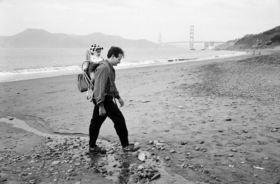 A man walks along a sandy beach carrying a child in a backpack carrier. The Golden Gate Bridge is visible in the background on a cloudy day.