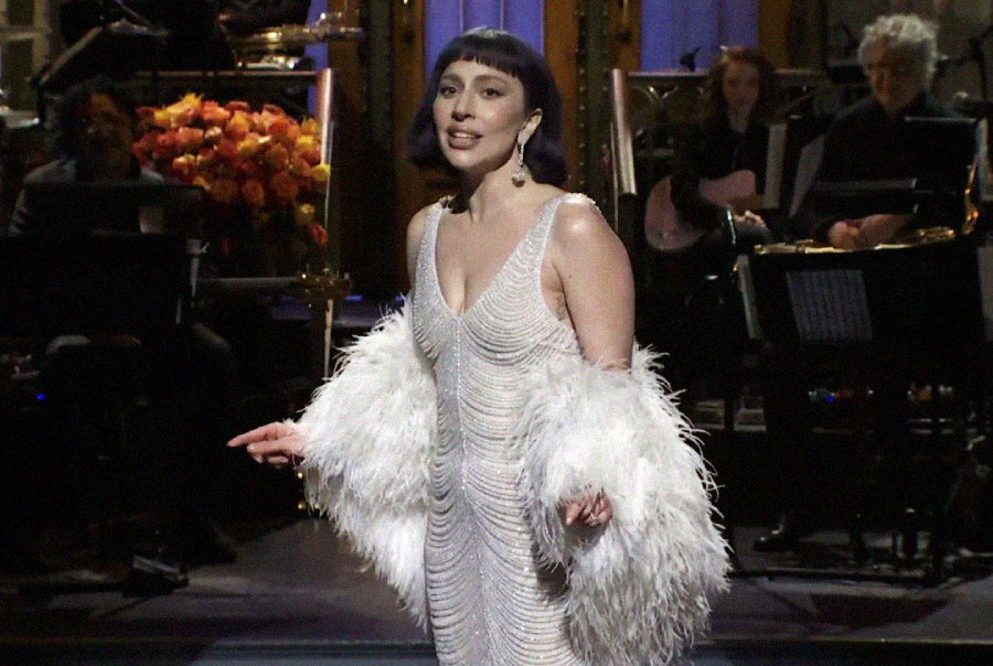 A woman in a sparkling white dress with a feathered shawl stands on stage, smiling and gesturing, with musicians seated and playing instruments in the background.