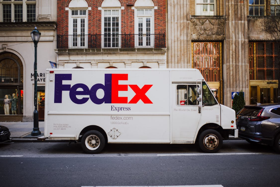 A white FedEx Express delivery truck is parked on a city street between buildings and other cars, with part of a sidewalk and several storefronts visible in the background.
