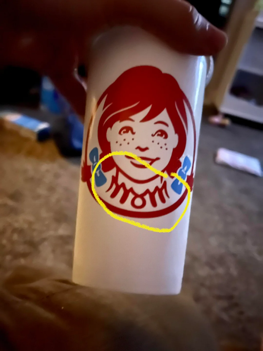 A hand holds a Wendy’s cup with the logo girl’s collar circled in yellow, highlighting that the collar spells “MOM” in its design. The background is a carpeted room with blurred objects.