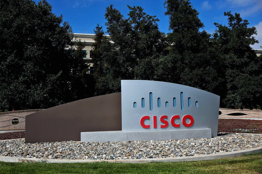 A large outdoor Cisco sign with the company logo in red and black sits on a landscaped area with rocks and grass, with trees and a building in the background.