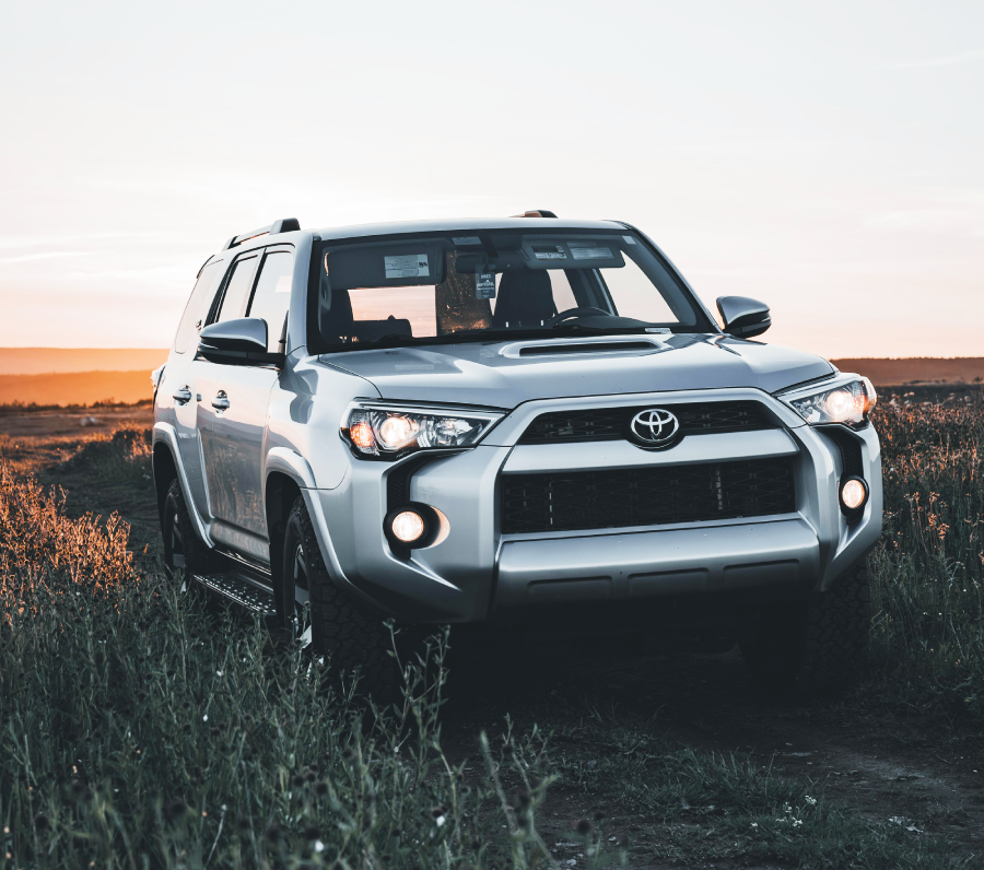 A silver Toyota SUV is parked on a grassy field at sunset, with its headlights on and tall grass surrounding the vehicle.
