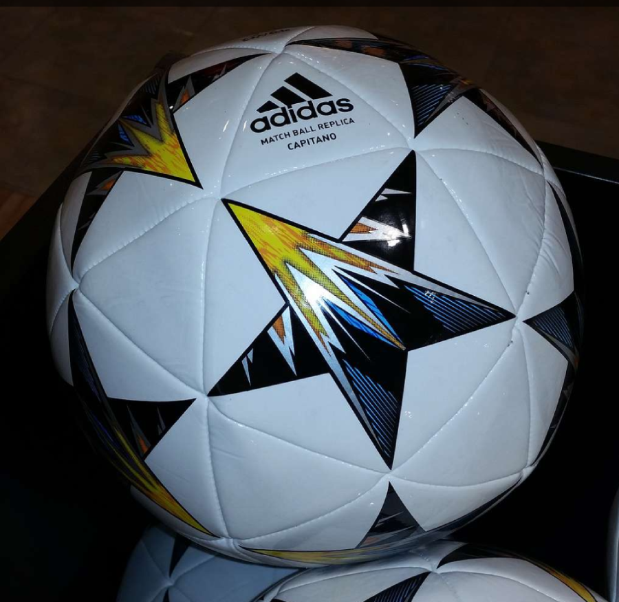 A close-up of an Adidas Match Ball Replica Capitano soccer ball with a white surface and black star patterns featuring yellow and orange accents. The Adidas logo and text are visible on the ball.