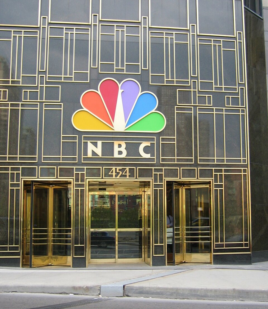 The entrance to an NBC building, featuring gold-framed glass doors and a large, colorful NBC peacock logo above the street number 454.