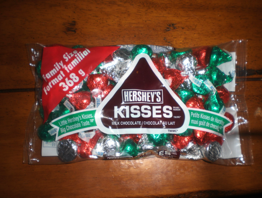 A 368g family size bag of Hershey’s Kisses milk chocolates in red, green, and silver foil wrappers sits on a wooden surface. The packaging is clear with brown and white labeling.