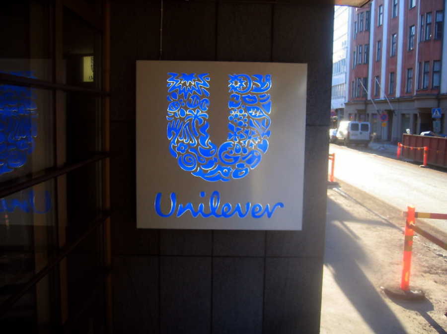 A sign with the Unilever logo and name is mounted on a building exterior. The logo is a large blue "U" made up of various icons, and sunlight is visible on the street beside the building.