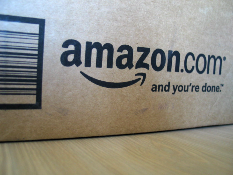 A close-up of a brown cardboard box with the Amazon.com logo and the slogan "and you're done." A barcode is partially visible on the left side of the box. The box rests on a wooden surface.