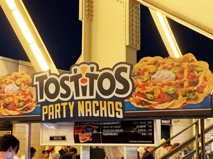 A brightly lit concession stand sign displays "Tostitos Party Nachos" with images of nachos topped with cheese, jalapeños, tomatoes, olives, and sour cream. People stand below the sign, and a menu is partly visible.
