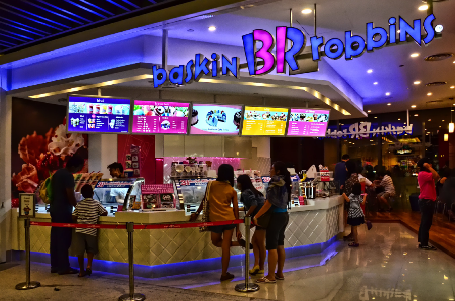 People stand in line at a brightly lit Baskin Robbins ice cream shop, with colorful menu boards and signs above the counter. The store has a modern design and a glowing purple and blue logo.