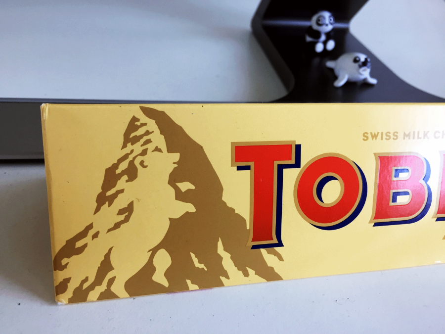 A close-up of a Toblerone chocolate bar box showing part of the logo and the mountain graphic. In the background, two small animal figurines sit on a black stand.