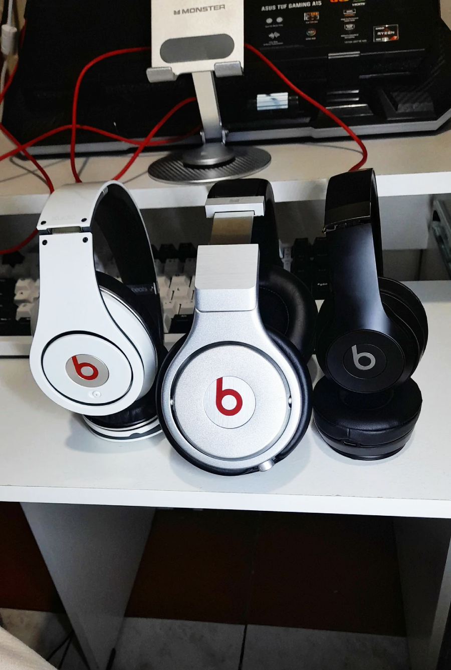 Three pairs of Beats headphones—one white, one silver, and one black—are displayed upright on a white desk, with a computer monitor and audio equipment visible in the background.