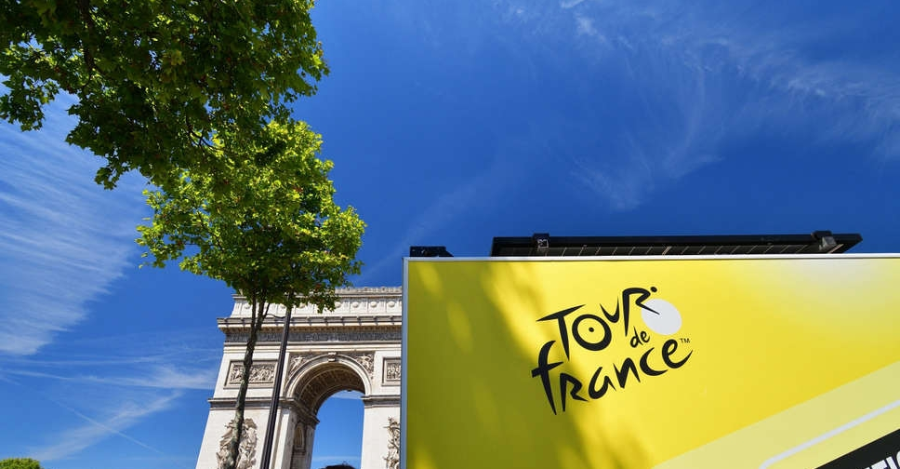 The Arc de Triomphe and a leafy tree are seen under a blue sky, with a large yellow Tour de France sign in the foreground.