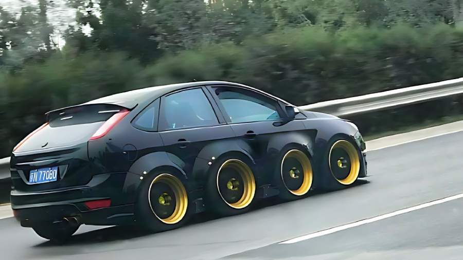 A black hatchback car with six wheels and gold rims is driving on a road, surrounded by greenery and a guardrail.