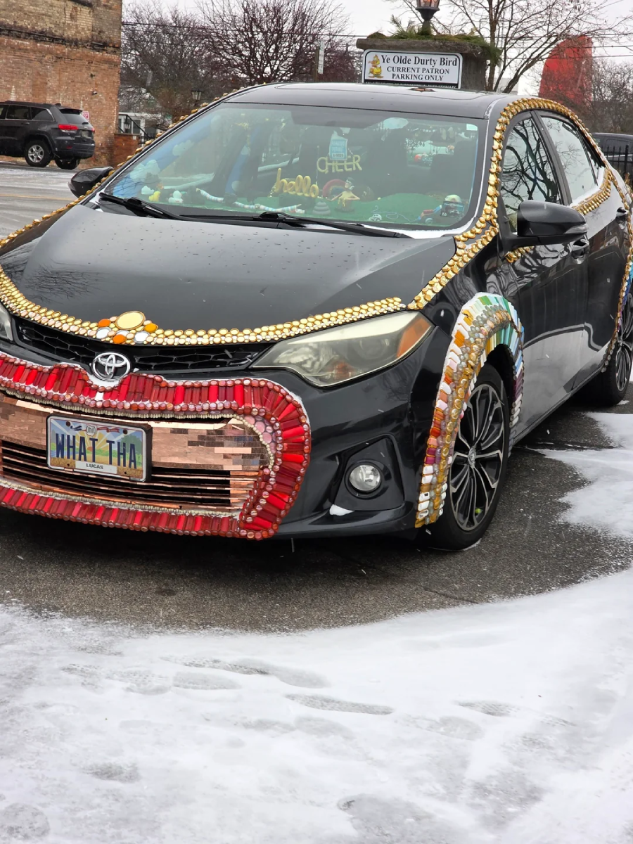 A black Toyota car decorated with colorful bottle caps, beads, and sequins, especially on the front and wheel arches. The license plate reads "WHAT THA." Snow is visible on the ground around the car.