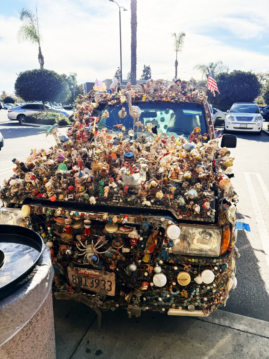 A car covered in hundreds of small toys, figurines, and trinkets, with American flags on the roof, parked in a sunny lot. The windshield and headlights are surrounded by decorations.