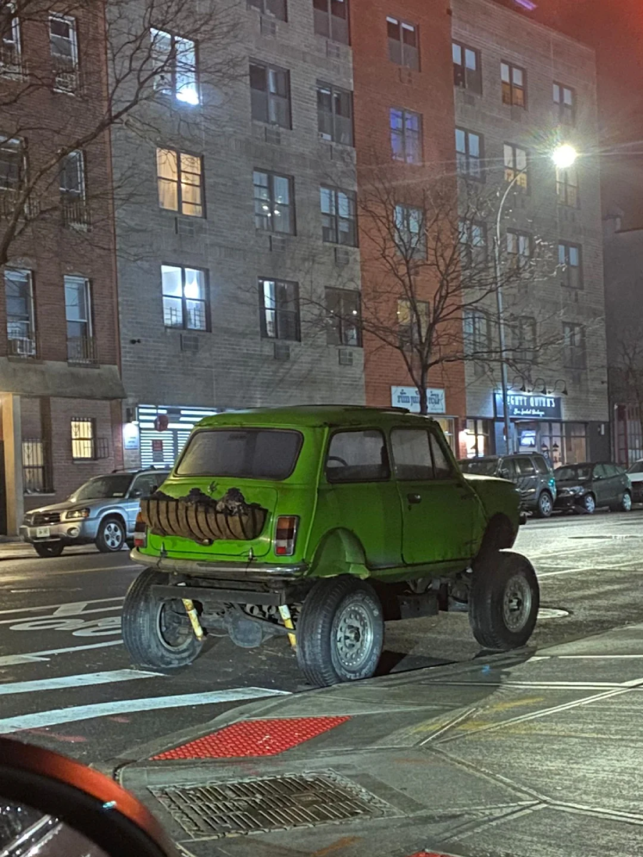 A small green car with very large off-road tires is parked on a city street at night. The car's rear features a spare tire and appears to be modified for extreme height. Apartment buildings and streetlights are in the background.