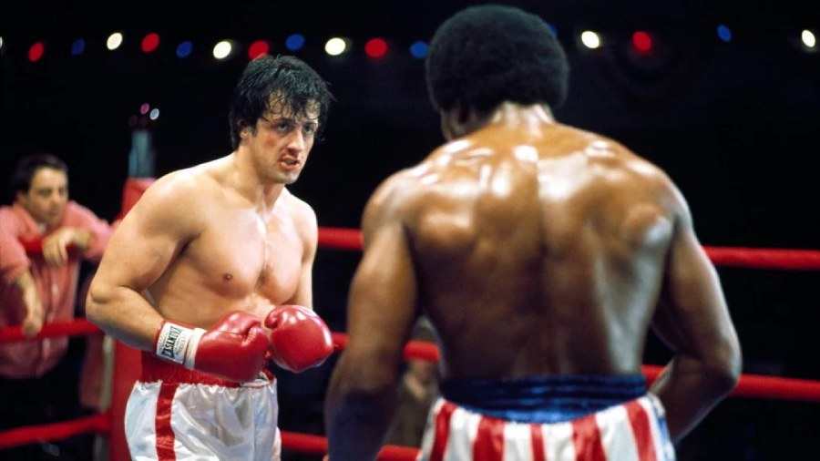 A boxer with short dark hair and red gloves faces an opponent with a muscular build and striped shorts in a boxing ring, under bright lights and watched by a blurred audience.