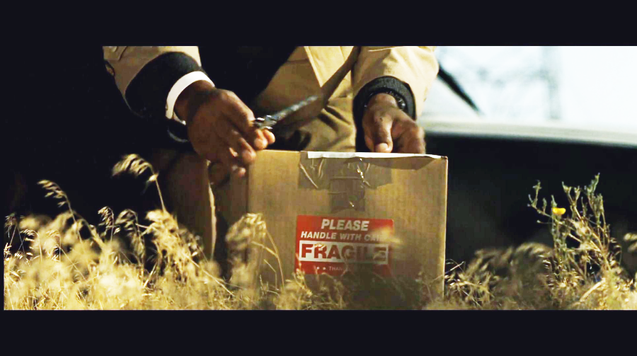 A person in a tan coat cuts the tape on a cardboard box labeled "PLEASE HANDLE WITH CARE FRAGILE" while kneeling in dry grass outdoors.