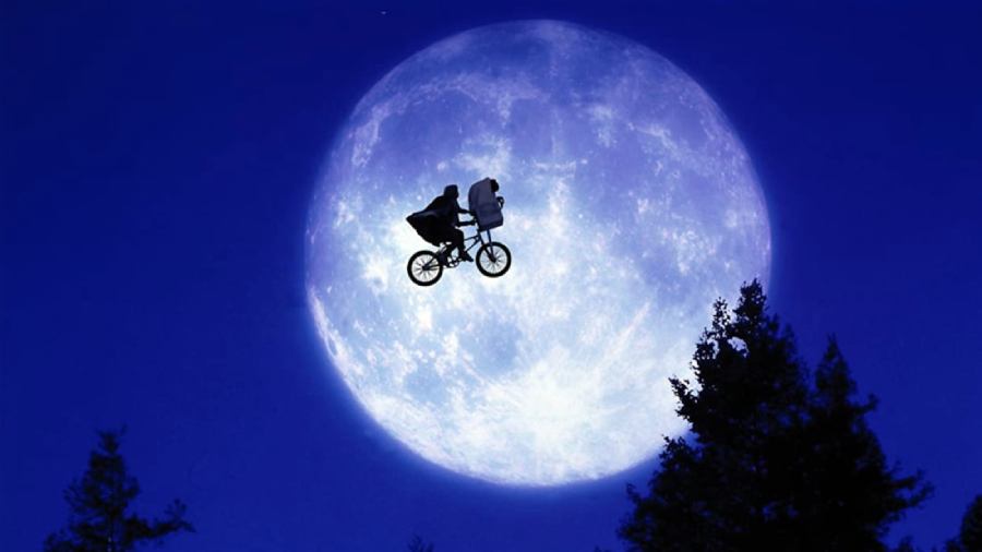 A person on a bicycle with a passenger in the front basket is silhouetted against a large, bright full moon in a dark blue night sky, with tree tops visible below.
