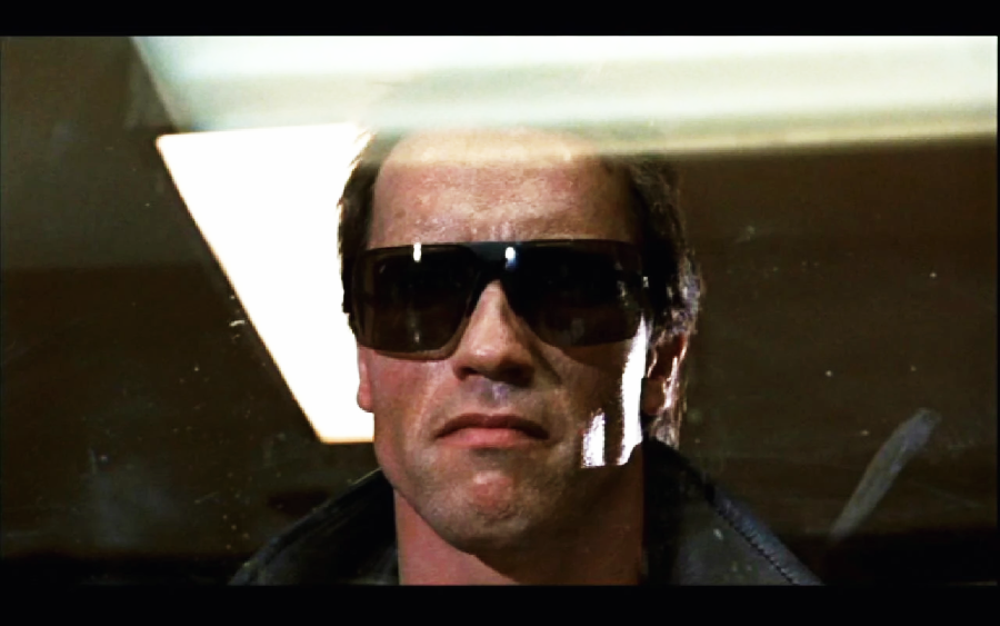 A man with a stern expression wears dark sunglasses and a black jacket, standing under a bright overhead light, with part of his face illuminated and the background mostly dark.