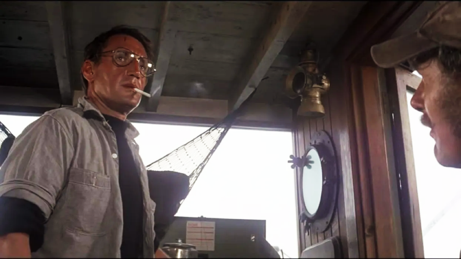 A man with glasses and a cigarette in his mouth stands on a boat, looking intently at another person. Nautical objects like ropes and a brass lamp hang in the background.