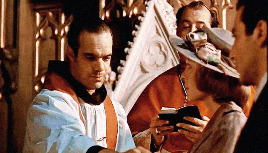A priest in white and red vestments stands next to another clergyman, holding a book, as he faces a woman in a hat and a man, appearing to conduct a wedding ceremony in a church.