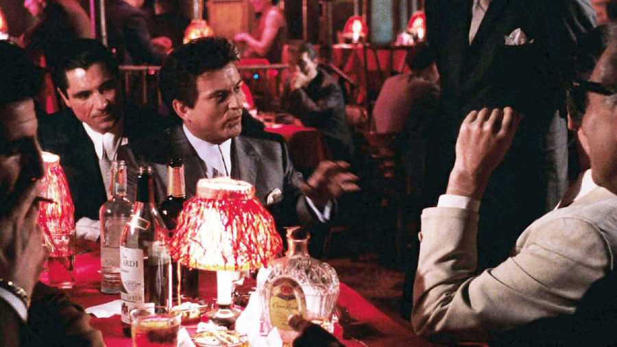 Four men in suits sit around a dimly lit table with red lamps, bottles, and glasses in a lively club or bar, engaged in conversation. The atmosphere is warm, crowded, and intimate.