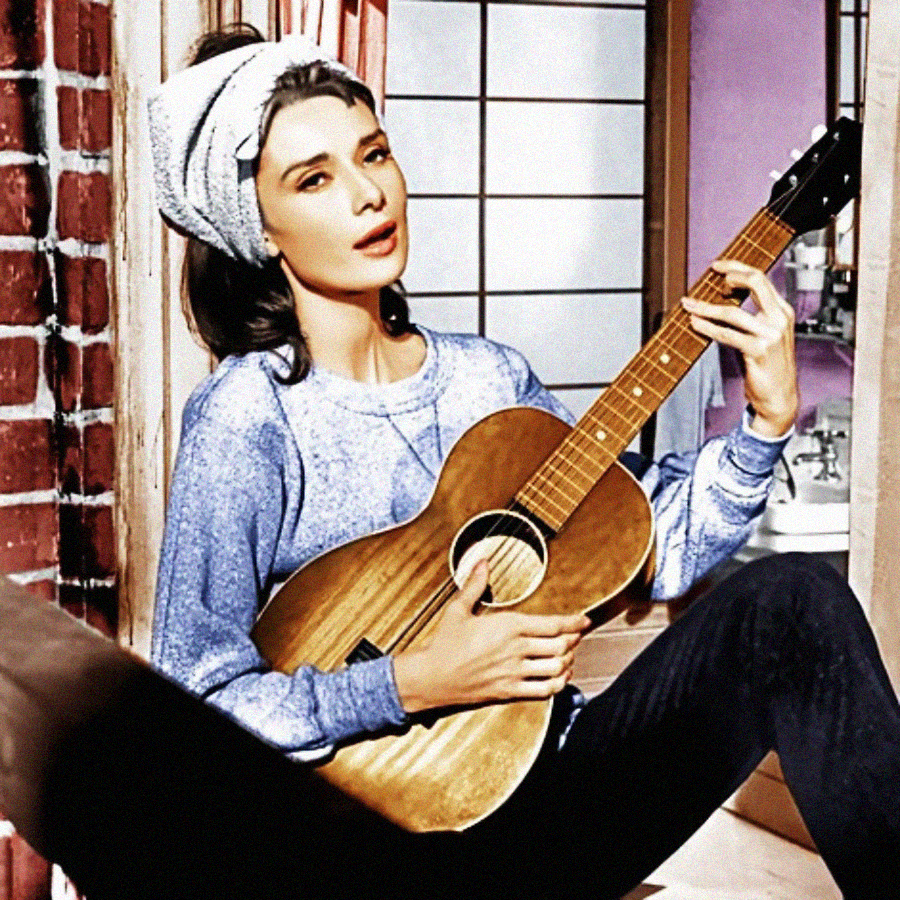 A woman with a towel wrapped around her head sits by a window, wearing a casual sweater and holding an acoustic guitar, strumming it as she looks thoughtfully into the distance.