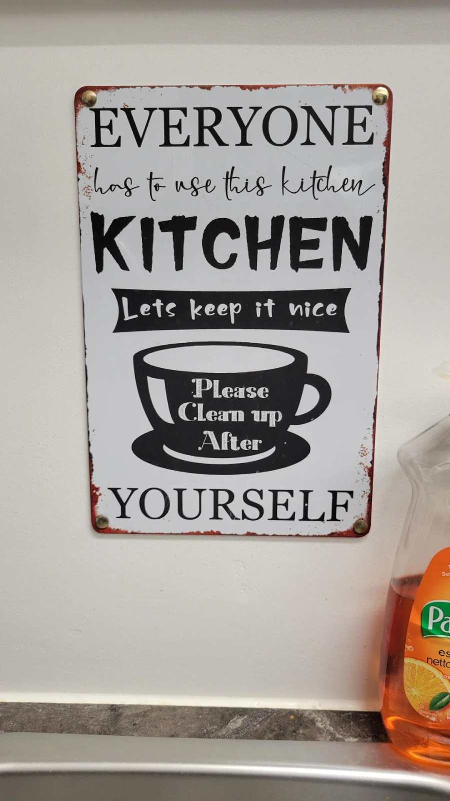 A sign on a white wall reads: "Everyone has to use this kitchen. Kitchen. Lets keep it nice. Please clean up after yourself." A bottle of Palmolive dish soap is visible on the right.