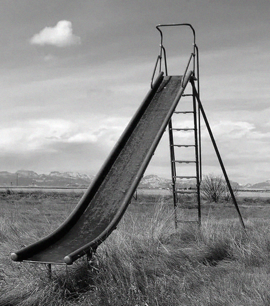 A tall, metal playground slide stands alone in an open, grassy field with mountains and clouds in the background. The image is in black and white, giving it a nostalgic, deserted feeling.