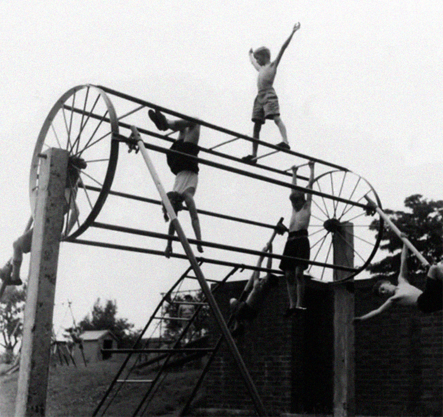 Black and white photo of children climbing and hanging from a tall, old-fashioned metal playground structure, some with arms raised, others dangling, surrounded by trees and brick walls in the background.