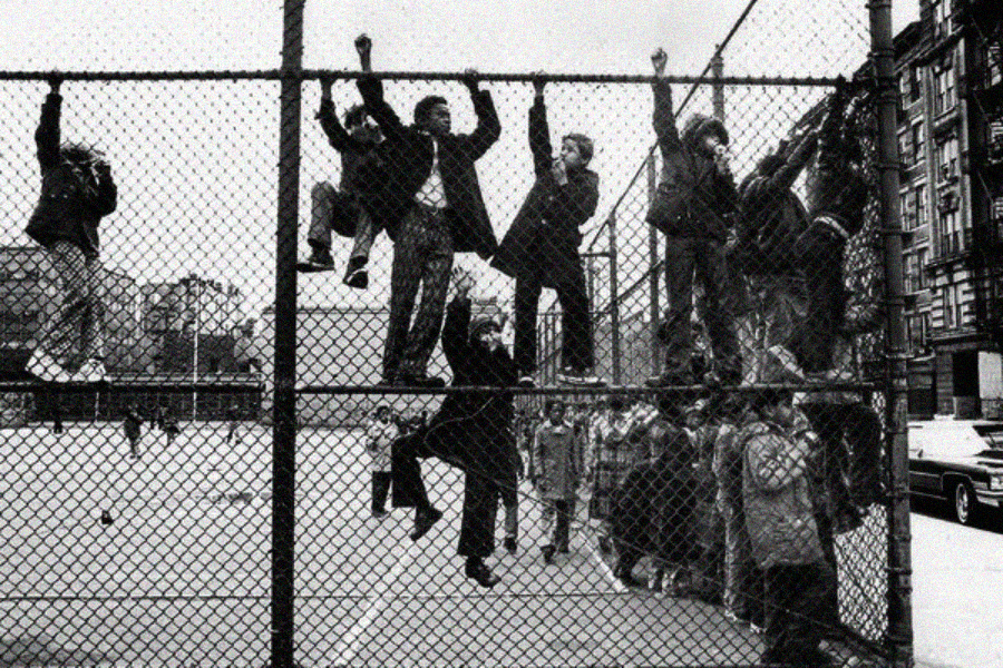 Children climb and hang from a tall chain-link fence at the edge of a city playground, while others stand and watch from the ground. Buildings and a parked car are visible in the background. The photo is black and white.