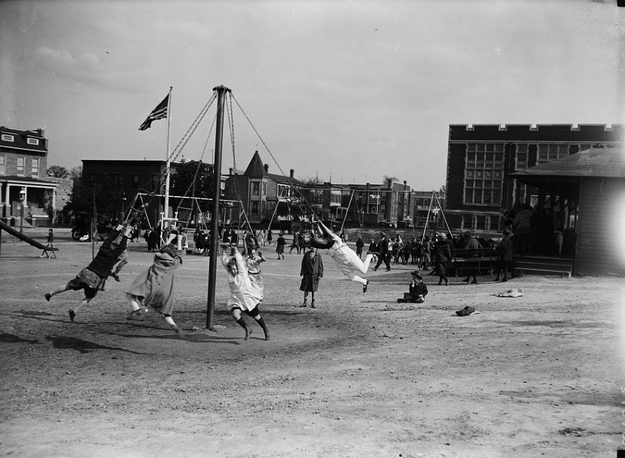 Children play on swings in a dirt playground, with more kids and adults in the background. Nearby are buildings, one with large windows, and a flagpole with a flag. The scene appears to be from the early 20th century.