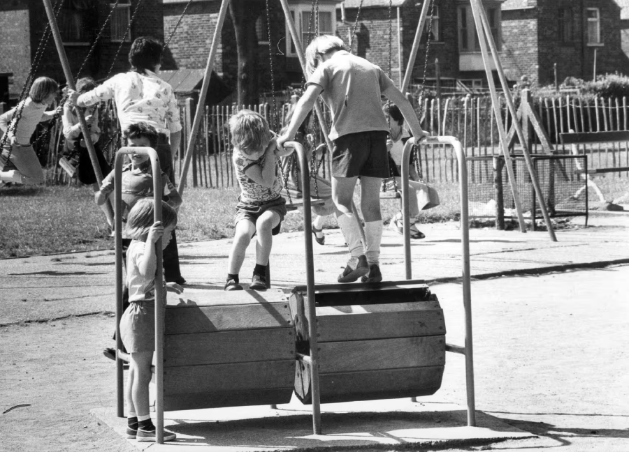 Black and white photo of children playing on a playground barrel climber and swings on a sunny day. Some kids are climbing while others watch or play in the background near a fenced grassy area.